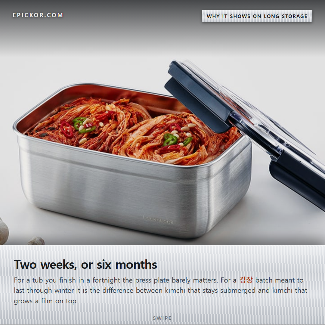 An open brushed stainless kimchi container packed with whole cabbage kimchi, its clear lid propped beside it and garlic on the counter