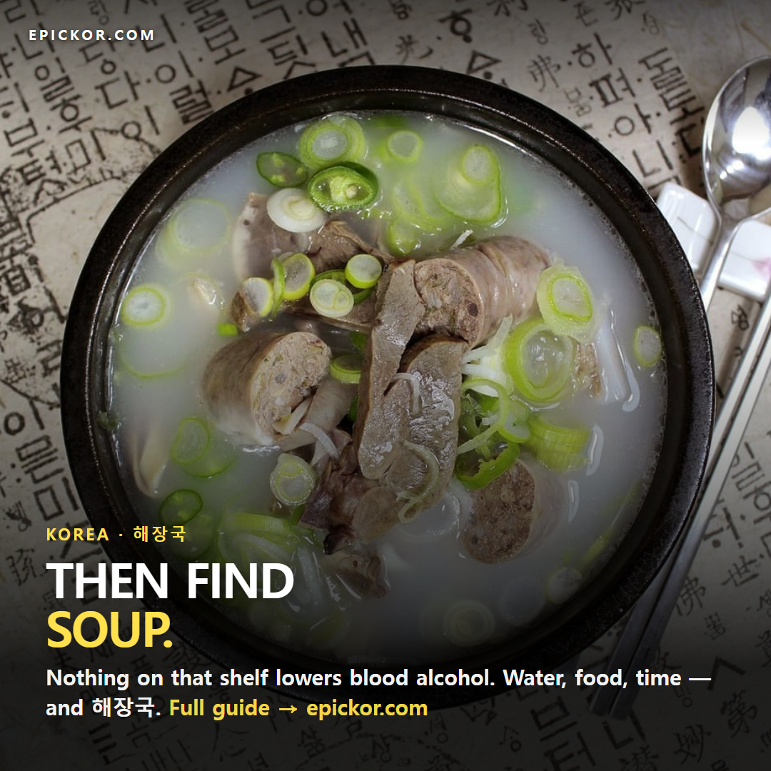 Overhead shot of a black stone bowl of Korean haejangguk (hangover soup) — milky broth with sliced meat, sundae and green onion — on Hangul-printed paper beside a spoon and chopsticks