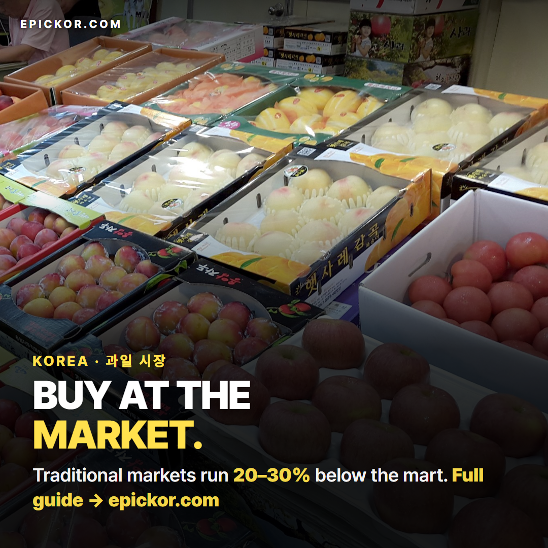 A Korean fruit market floor, July 2017 — a tray of red apples in front, boxes of peaches (햇사레 감곡) and plums behind, in Korean-labelled cartons (brightened derivative, gamma 0.68, contrast +8%)