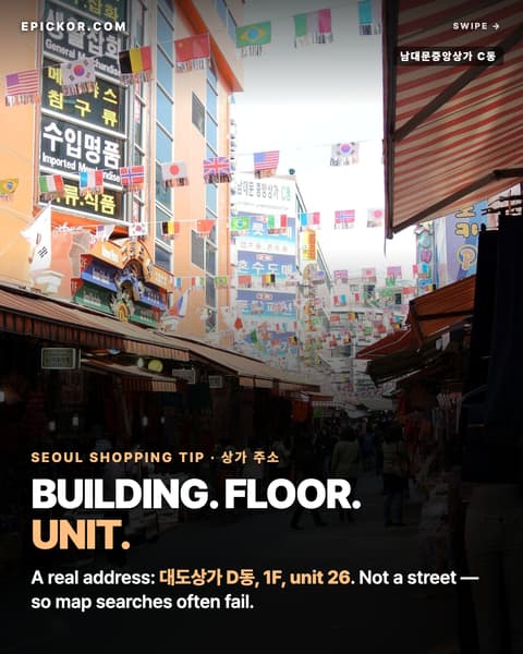 An alley inside Namdaemun Market under strung flags, with the large sign of 남대문중앙상가 C동 (Namdaemun Jungang Shopping Town, Building C) and bag and import shops on both sides (Adbar, Wikimedia Commons)
