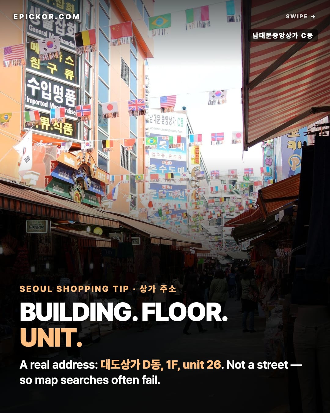 An alley inside Namdaemun Market under strung flags, with the large sign of 남대문중앙상가 C동 (Namdaemun Jungang Shopping Town, Building C) and bag and import shops on both sides (Adbar, Wikimedia Commons)