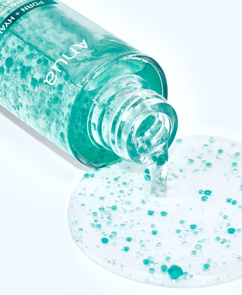 A tilted Anua PDRN capsule mist bottle pouring clear liquid with small turquoise capsules into a pool on a white surface.