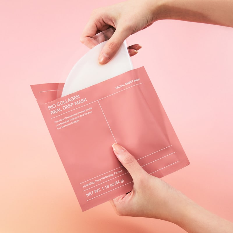 Two hands pulling a white hydrogel mask out of a pink Biodance Bio Collagen Real Deep Mask pouch, against a pink background.