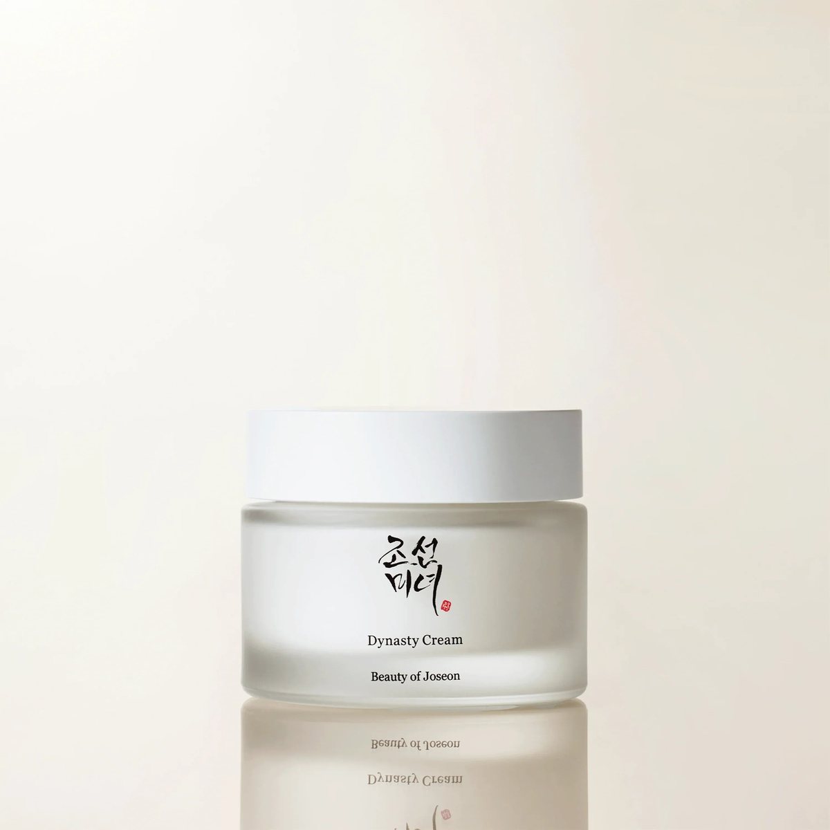 A frosted glass jar of Beauty of Joseon Dynasty Cream with a white lid and the brand's Hangul calligraphy logo, reflected on a glossy surface.