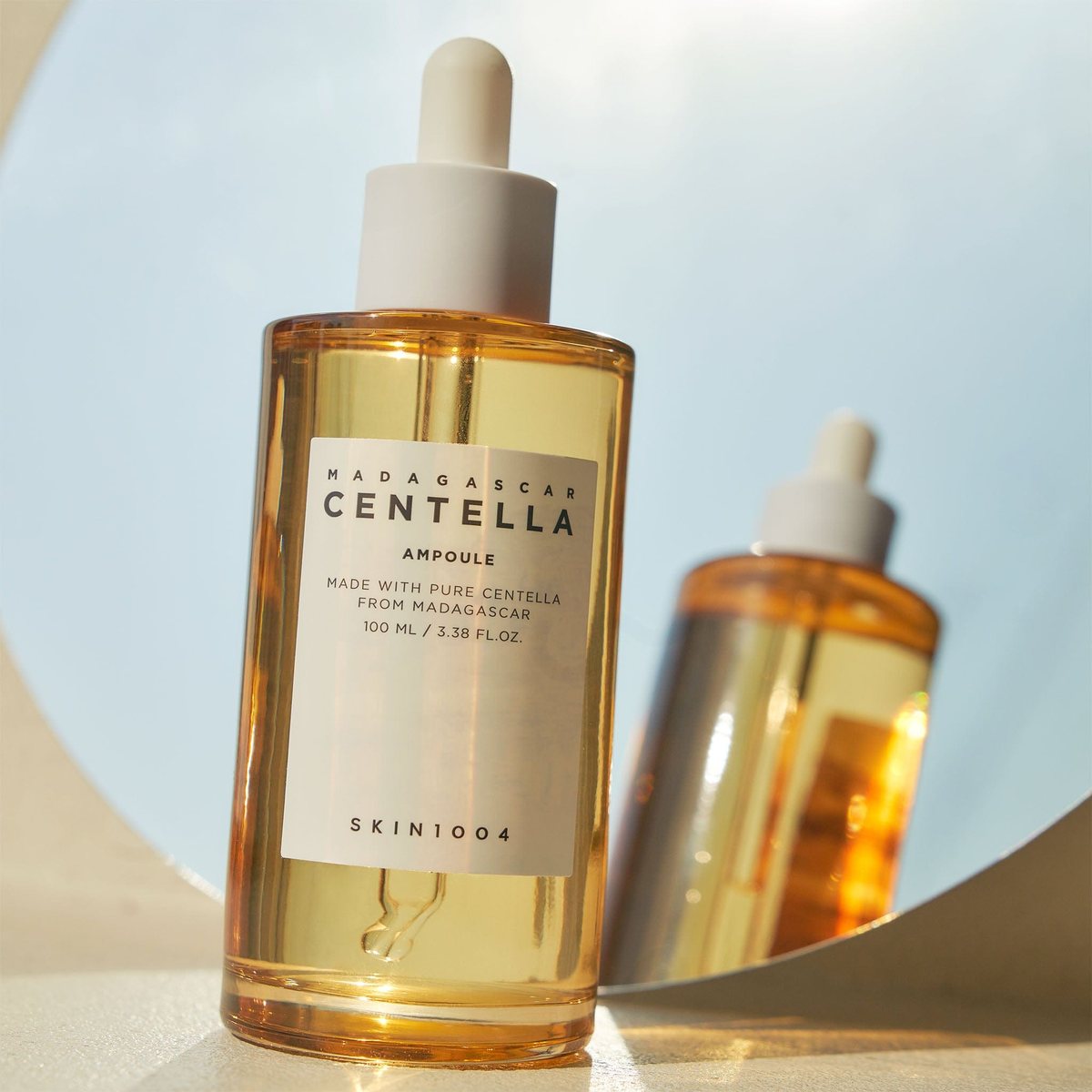 A bottle of SKIN1004 Madagascar Centella Ampoule with a white dropper, amber liquid and a white label, reflected in a round mirror.