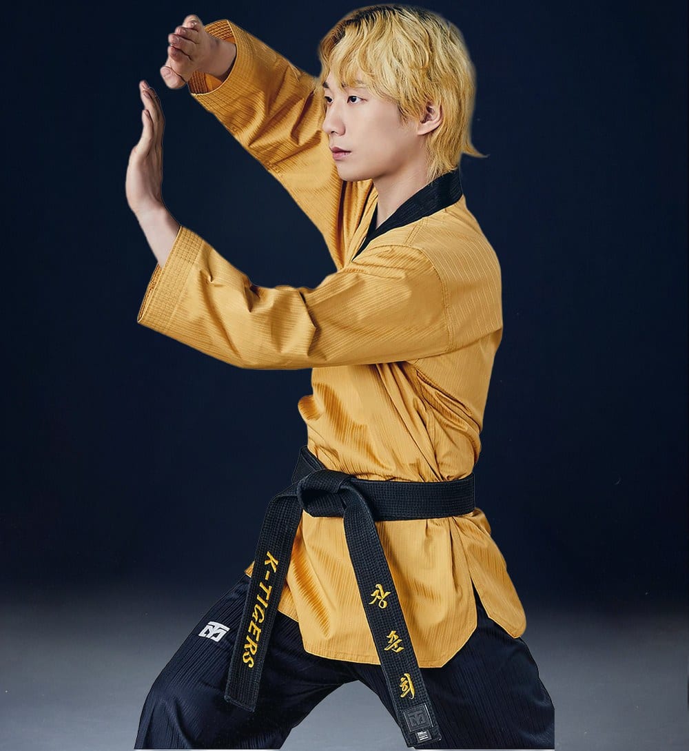 A taekwondo practitioner in a gold Mooto TAEBEK 3 poomsae uniform with a black collar and navy trousers, holding a poomsae stance against a dark blue background, wearing a black belt embroidered K-TIGERS.