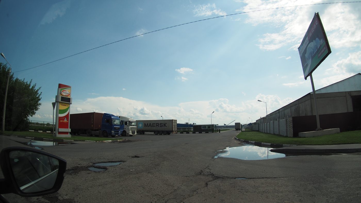 The entrance road to an industrial site in Ramenskoye, Moscow region, with a tall red, yellow and green Doshirak sign on the left, trucks and a Maersk container lorry parked by the gate, and low warehouse buildings on the right.