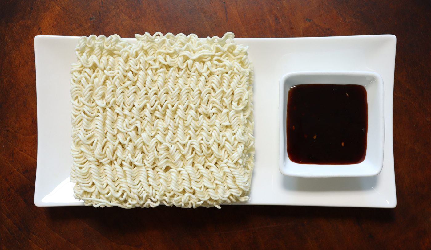 A square block of dry Paldo Bibimmen noodles on a white rectangular plate, next to a small square dish of dark red-brown bibim sauce.