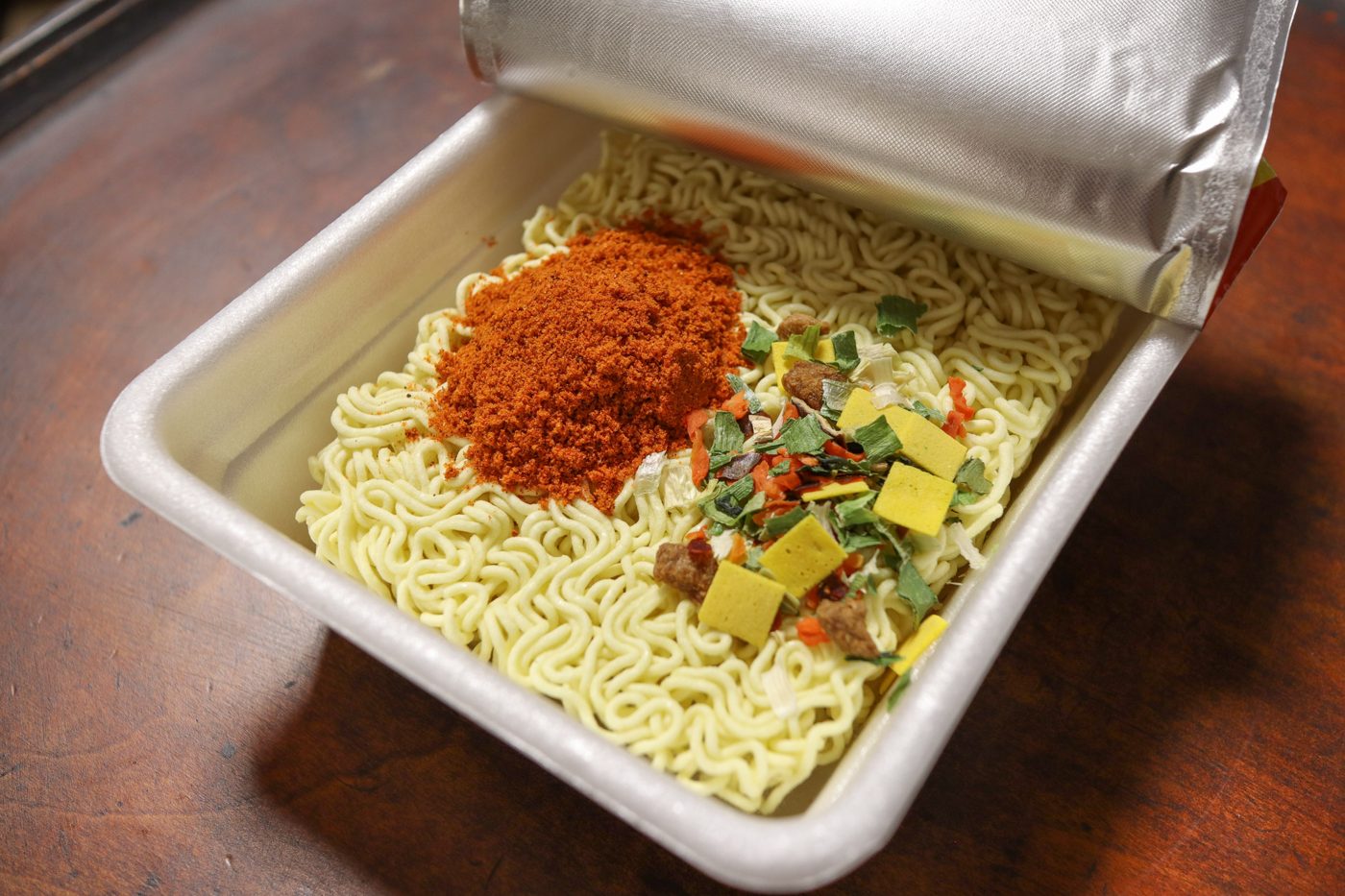 An opened Korean-market Paldo Dosirac: a square white tray of dry wavy noodles with a mound of red-orange soup powder and dried vegetable and egg flakes, the foil lid peeled back.