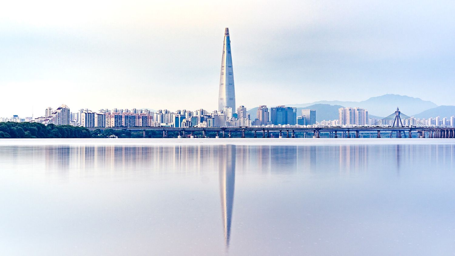 The Lotte World Tower in Seoul rising above apartment blocks and a bridge, reflected in the still Han River on a pale morning.