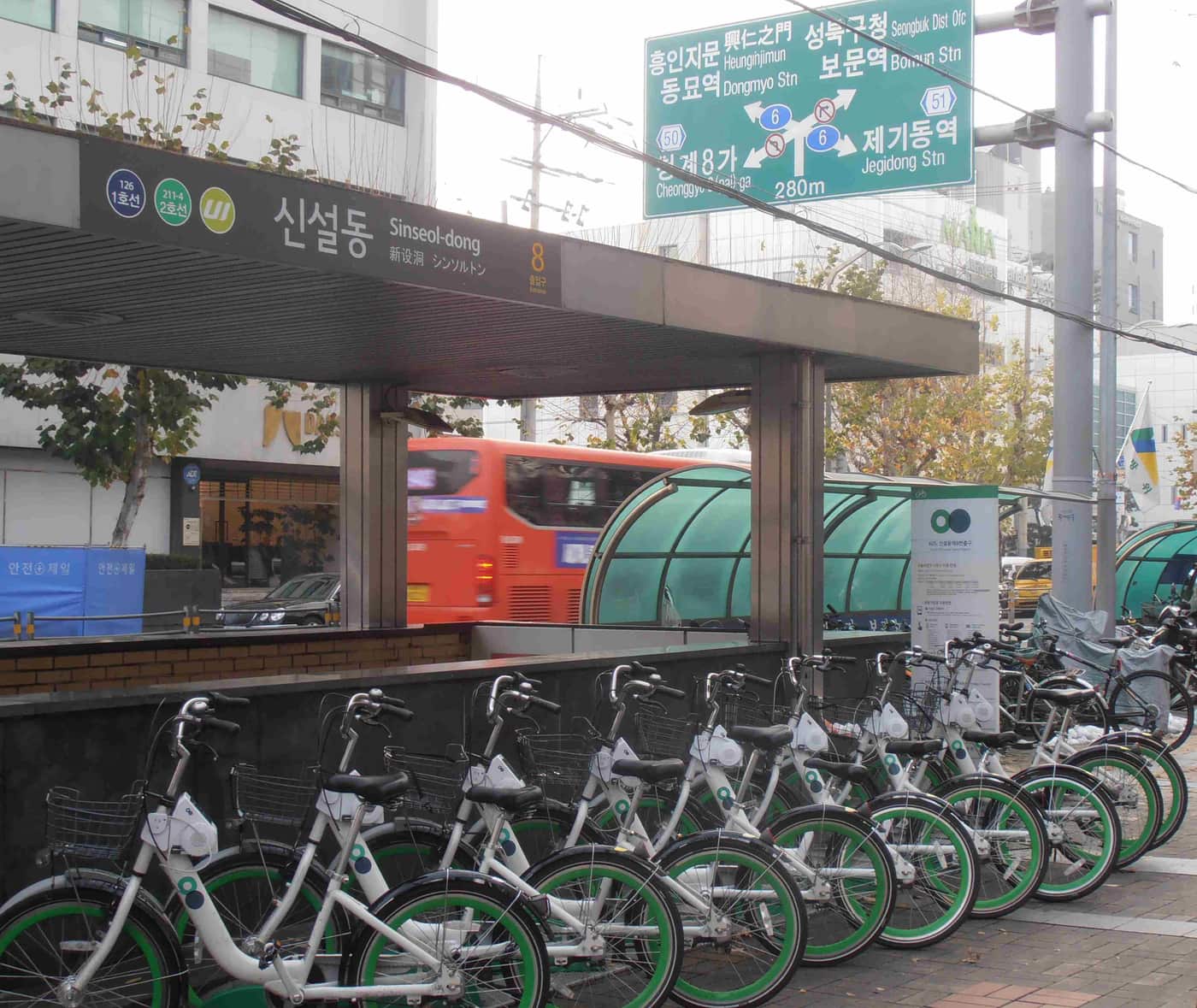 A rack of green-and-white Seoul Bike public hire bicycles parked outside Sinseol-dong Station exit 8, under Korean subway and road signage.