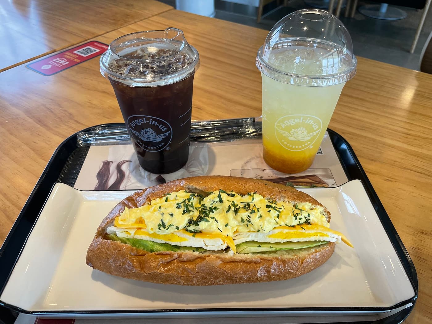 An iced americano in a takeaway cup at a Korean cafe, beside a sandwich and an ade.
