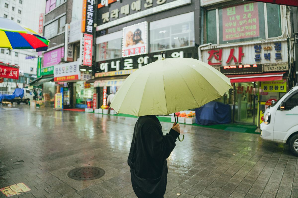 A rainy Seoul market street with umbrellas and Korean urban signs.