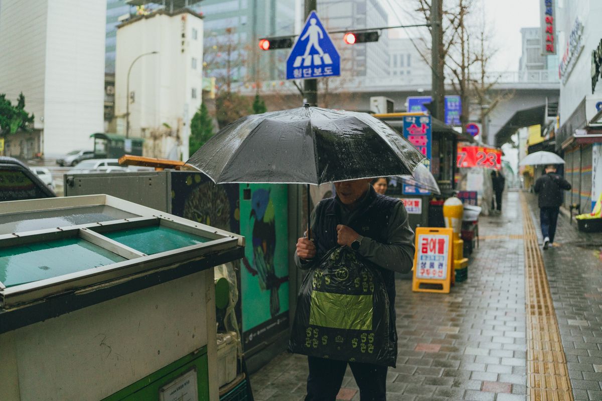 A rainy Seoul street with a person walking under an umbrella.