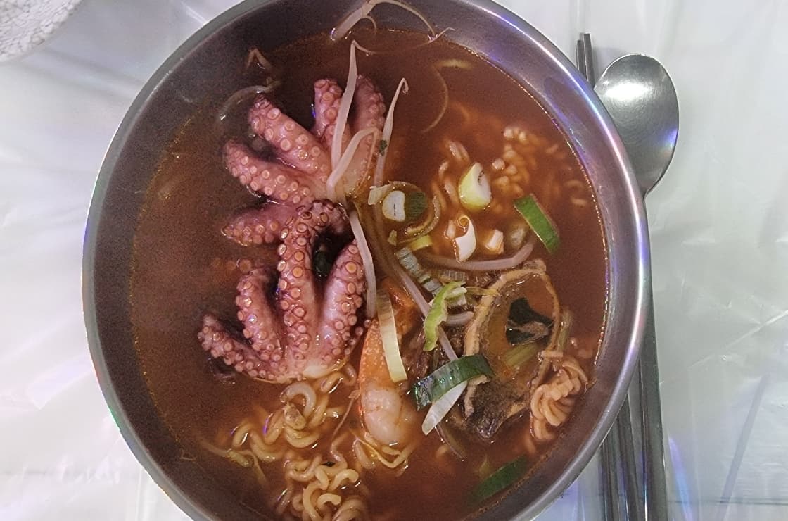 Seafood ramyeon in a stainless bowl with octopus, spring onion and a Korean spoon and chopsticks.