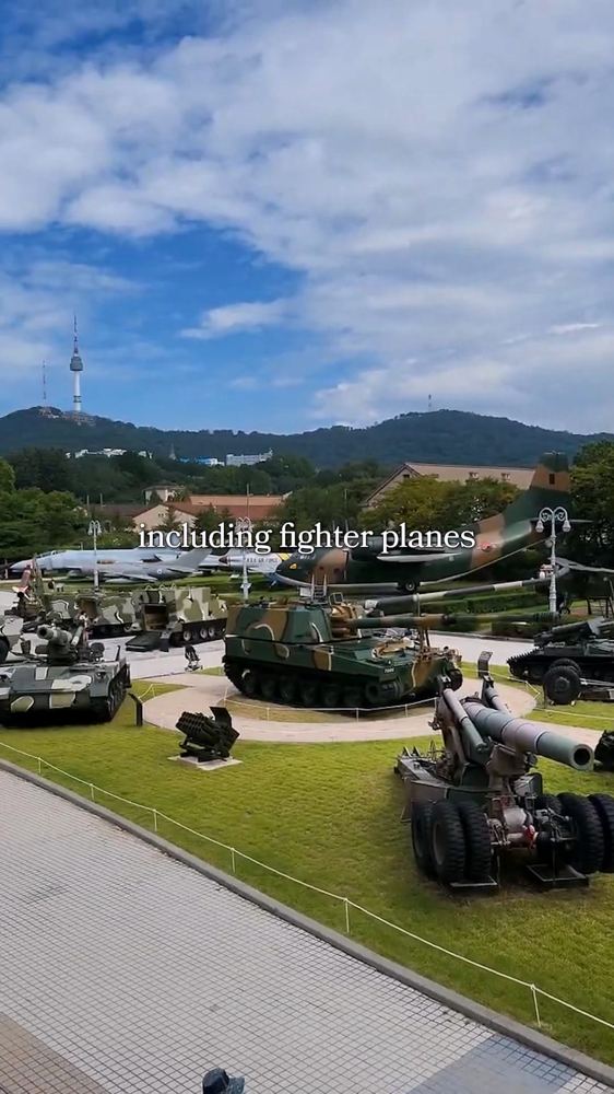 The outdoor exhibition at the War Memorial of Korea in Yongsan, Seoul: camouflaged tanks, howitzers and a transport aircraft on lawns, with N Seoul Tower on Namsan in the background.