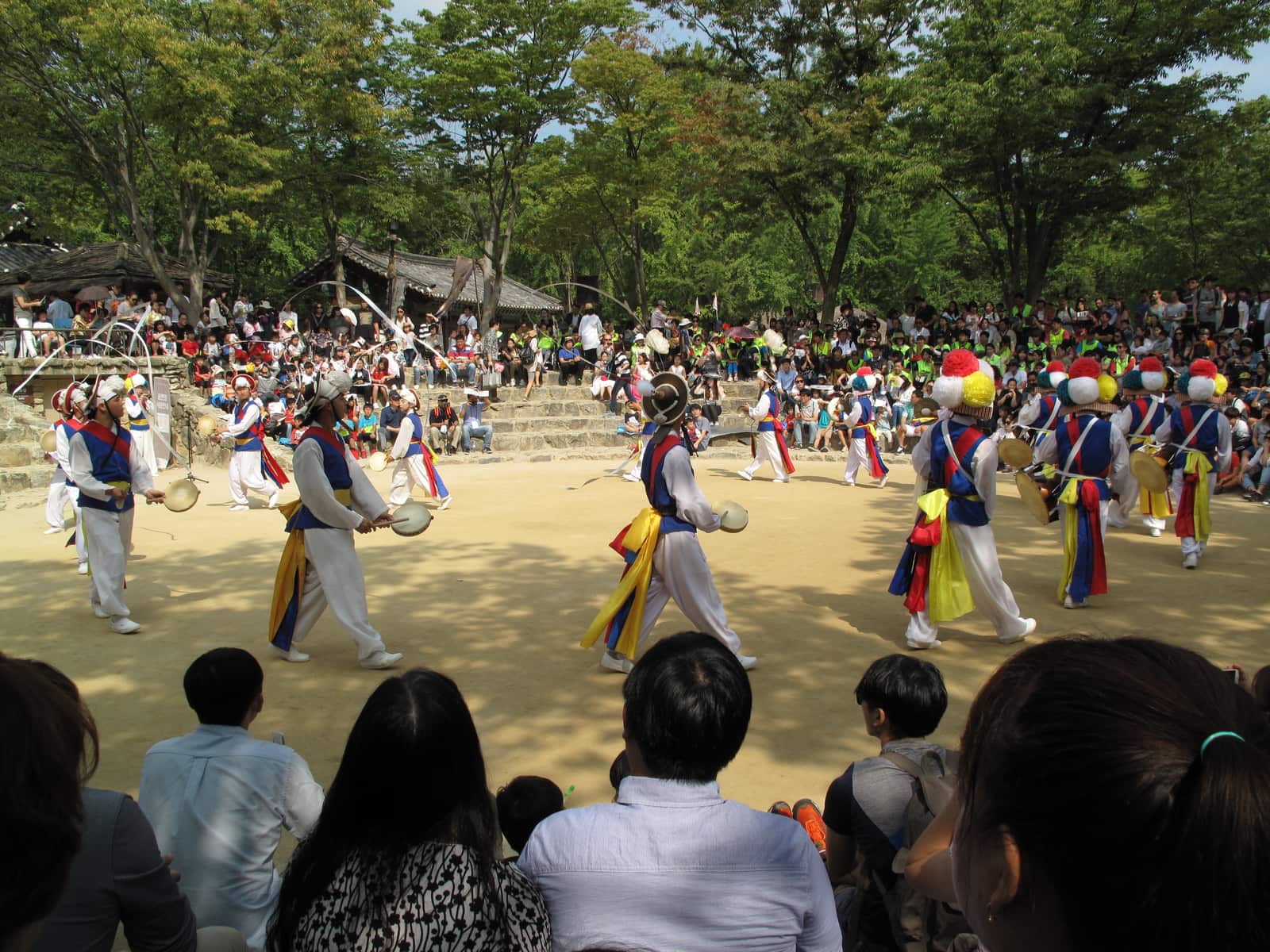 Pungmul farmers' dancers in blue vests and yellow sashes circling the sand arena at the Korean Folk Village, with a seated crowd on stone steps around them.