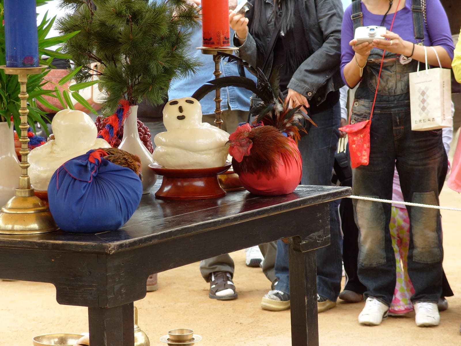 A ritual table for a traditional Korean wedding at the Korean Folk Village, with rice-cake figures, pine branches, and a rooster and hen wrapped in red and blue cloth, as visitors photograph it.