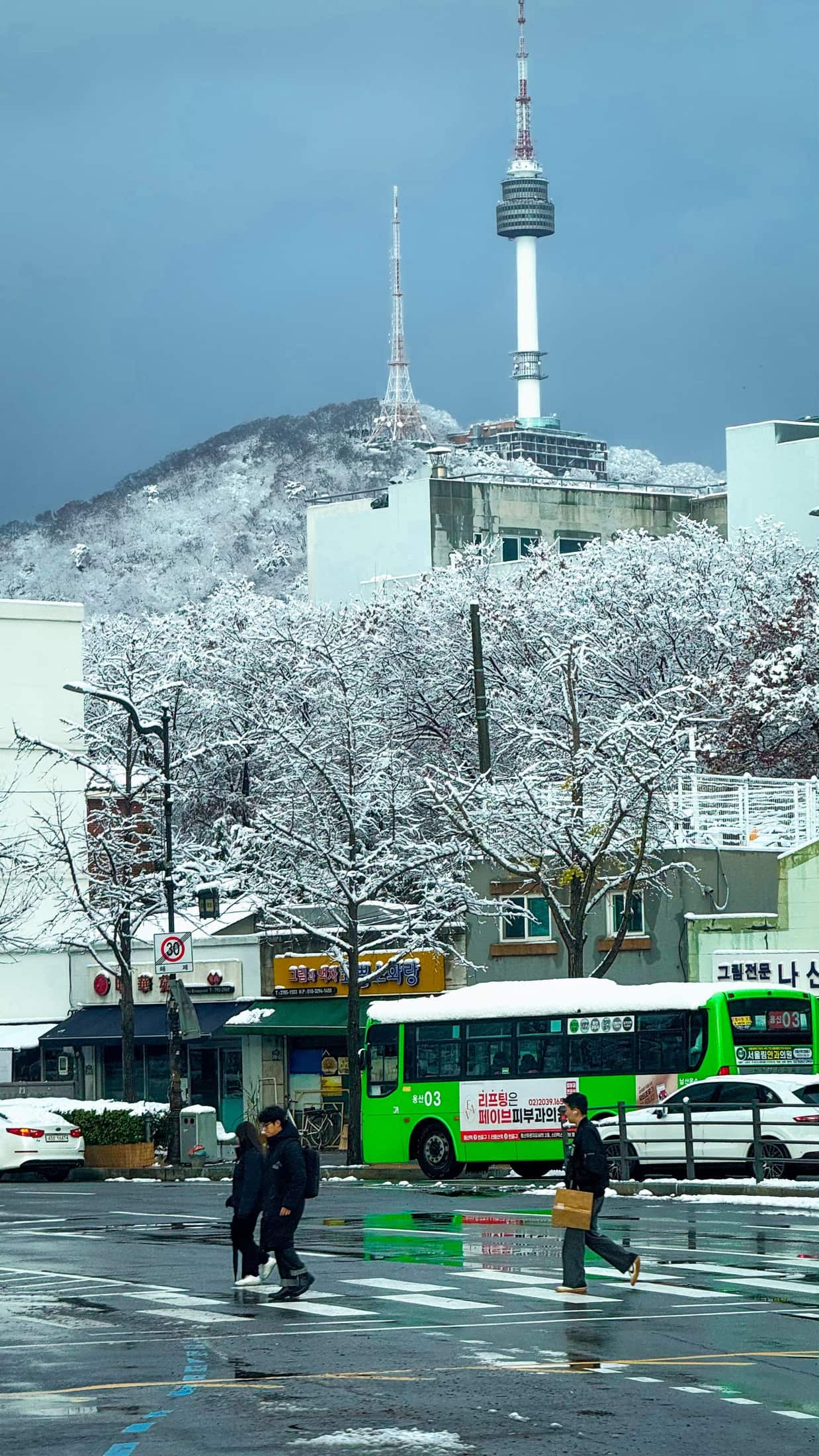 Snow-covered street below N Seoul Tower with pedestrians in dark winter coats crossing the road.