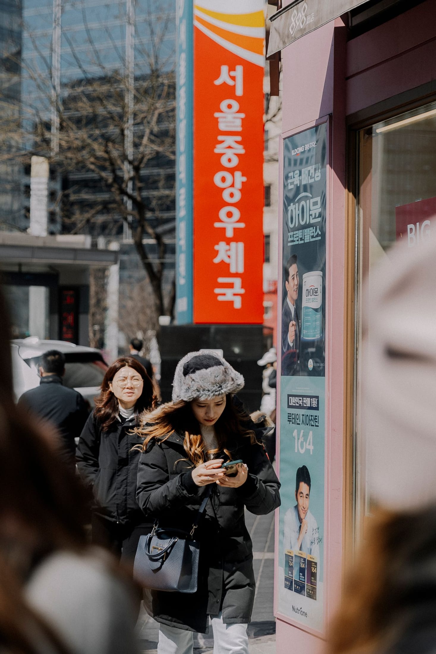 Two women in long black padded coats walking past a Seoul Central Post Office banner in winter.