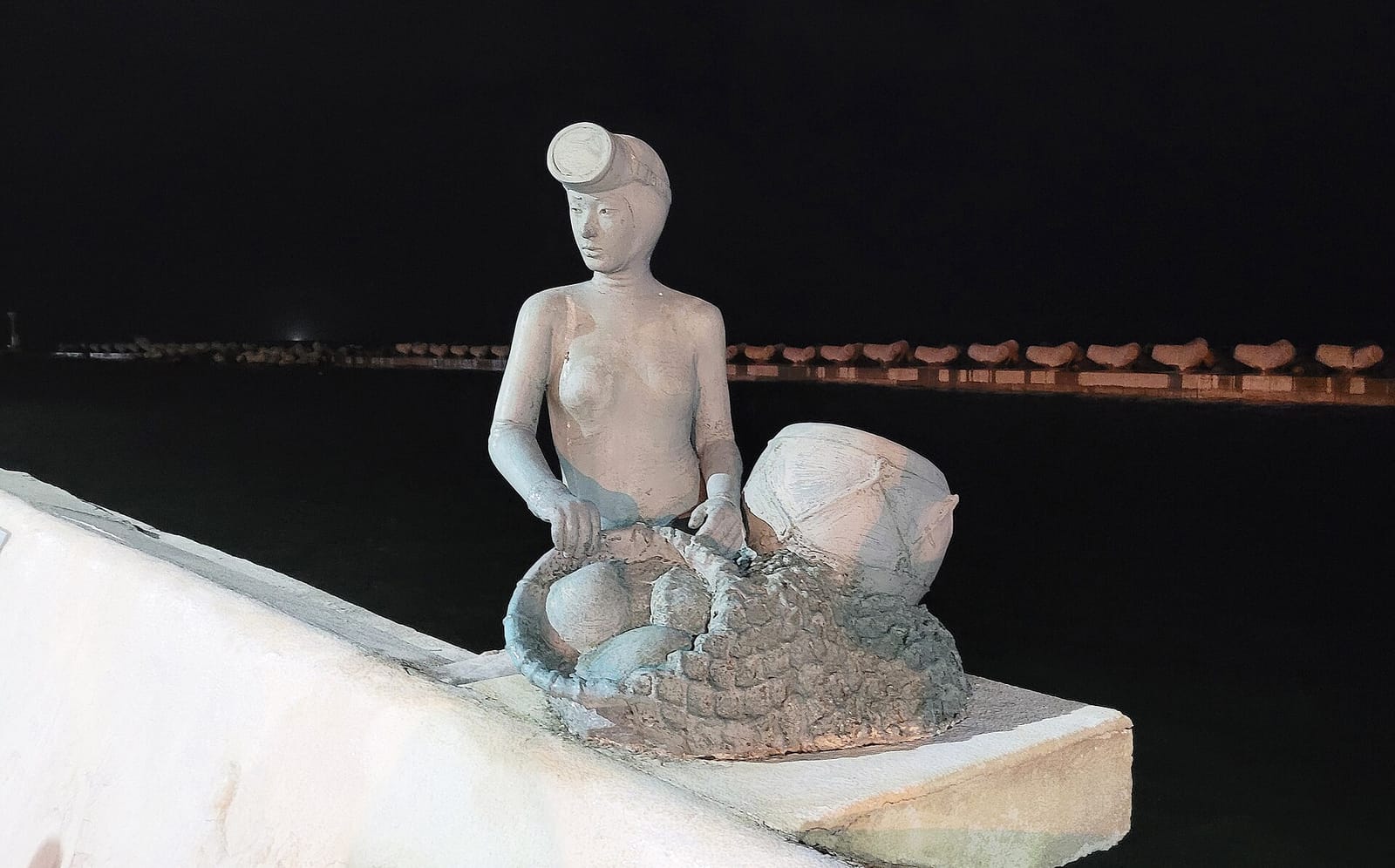 A stone statue of a haenyeo with her tewak and net on the Jeju coast at night.