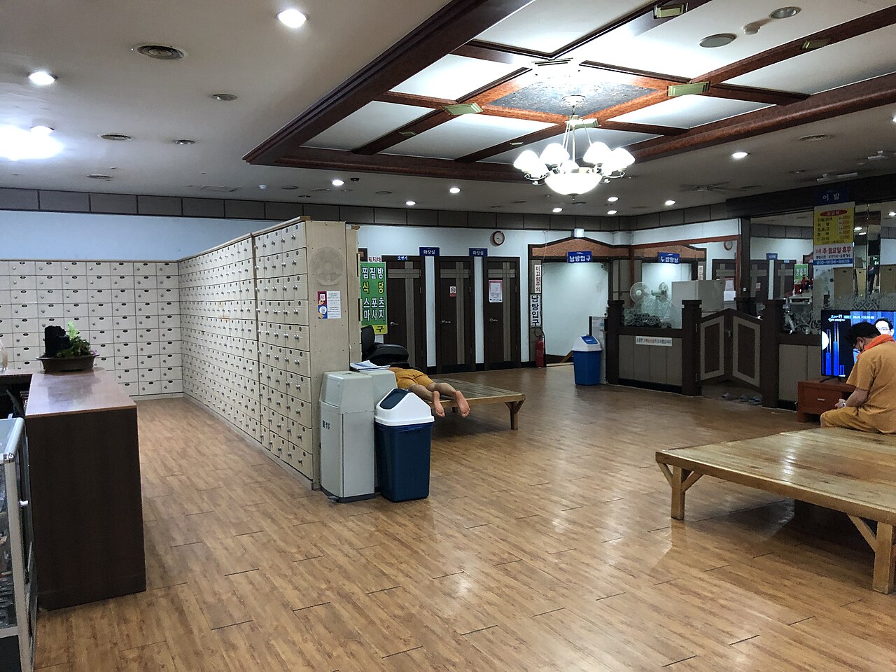 A Korean jjimjilbang locker and bathhouse area with rows of lockers, benches, and local facility signs.