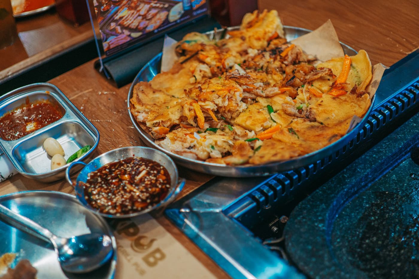 A large seafood pajeon on a steel plate at a Korean pub table, with a soy-vinegar dipping sauce, a divided steel dish of red sauce and garlic, and a built-in grill beside it.