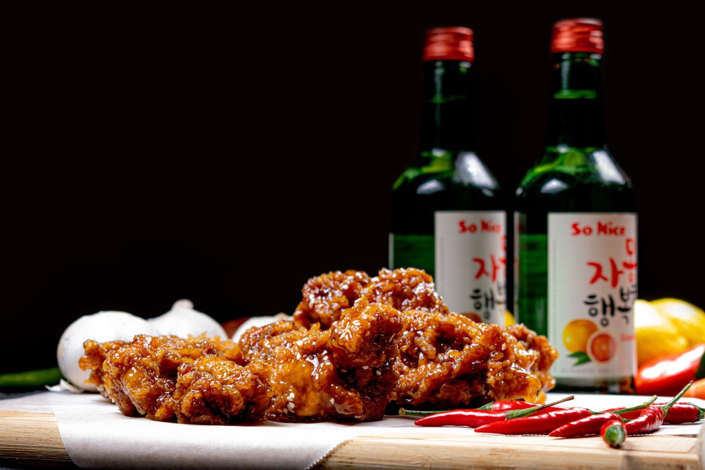 Korean fried chicken glazed in soy-garlic sauce on parchment, with two green bottles of grapefruit-flavoured soju, garlic, lemon and red chillies behind it.