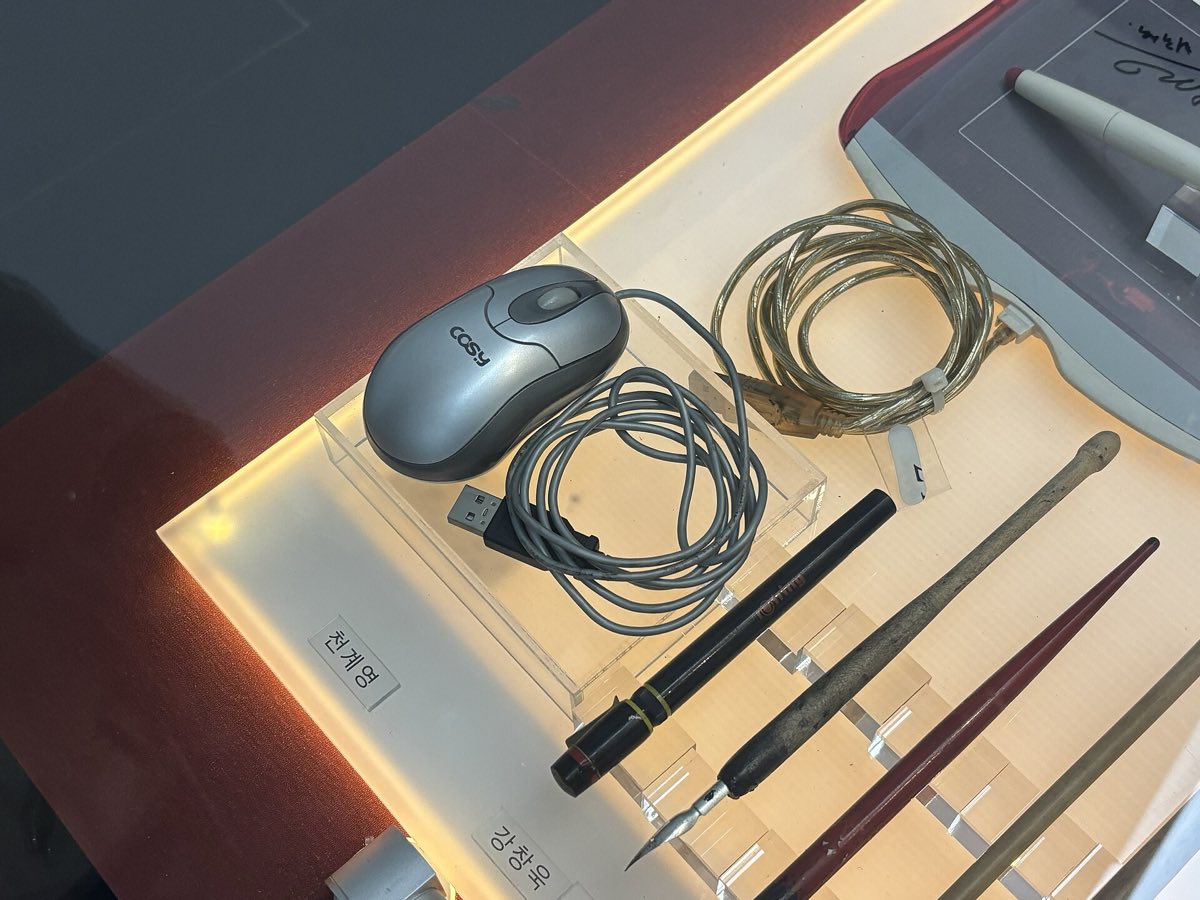 A display case at the Korea Manhwa Museum with dip pens, a computer mouse and a pen tablet laid side by side, with Korean name labels for the artists who used them.