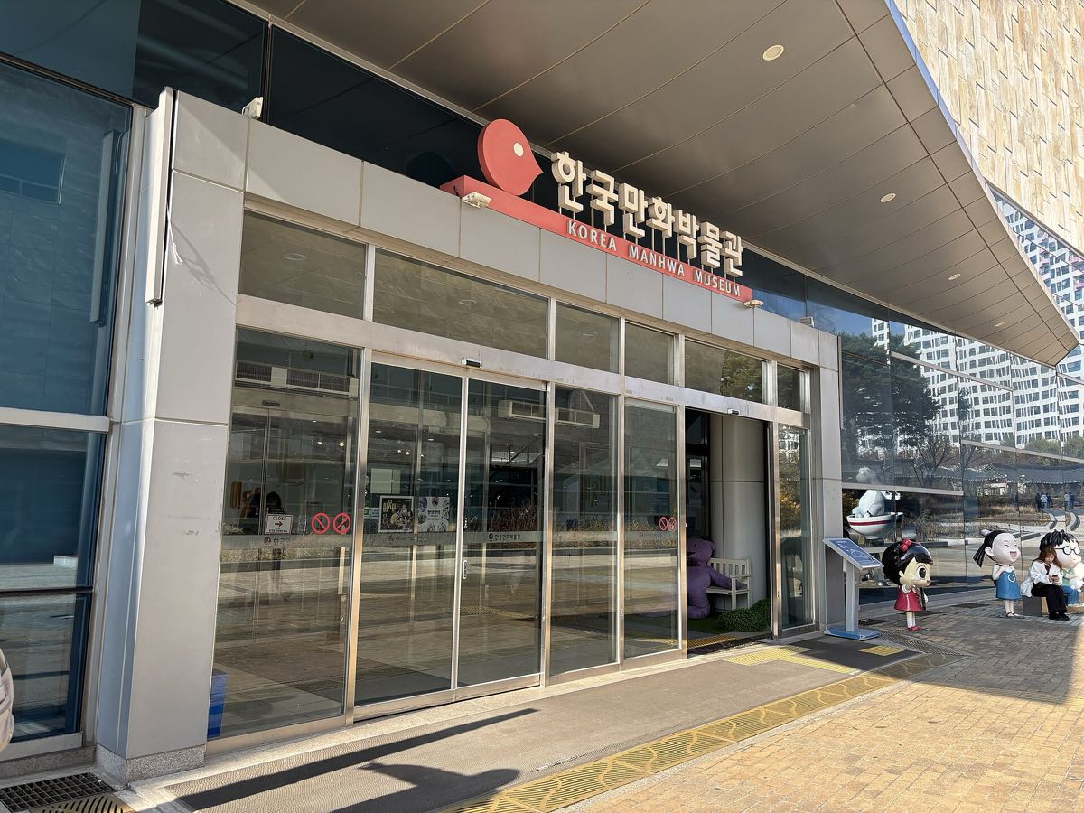 The entrance of the Korea Manhwa Museum in Bucheon, with the red 한국만화박물관 Korea Manhwa Museum sign above glass doors and cartoon character statues outside.