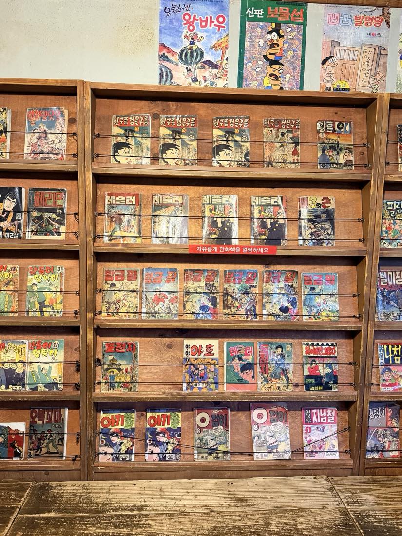 Older printed Korean manhwa on wooden shelves in the Korea Manhwa Museum's permanent exhibition, under a red sign that reads 'feel free to browse the comic books'.