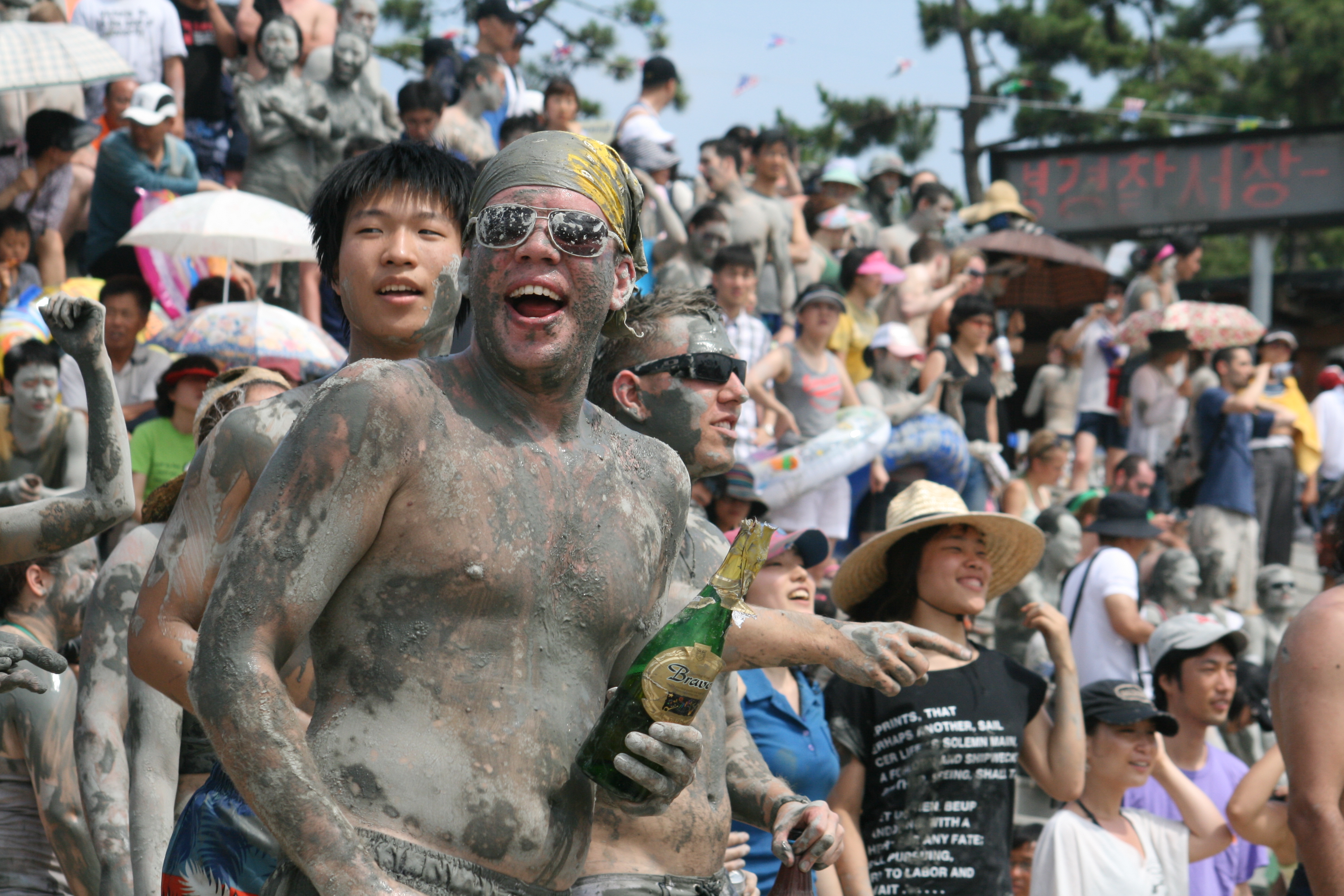 A mud-covered festivalgoer smiling in a crowded Boryeong Mud Festival activity area.