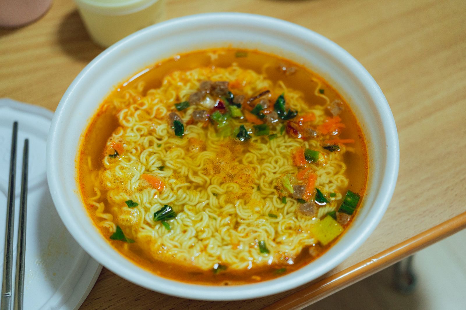 A close bowl of spicy Korean ramen with greens and broth, showing how add-ons turn instant noodles into a fuller meal.