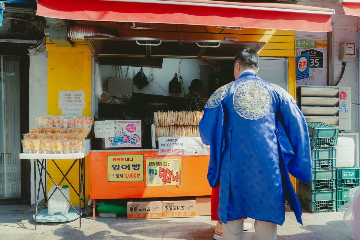 A Seoul food stall seen from behind a person in hanbok, showing how street snacks can overlap with tourist routes and cultural walking areas.