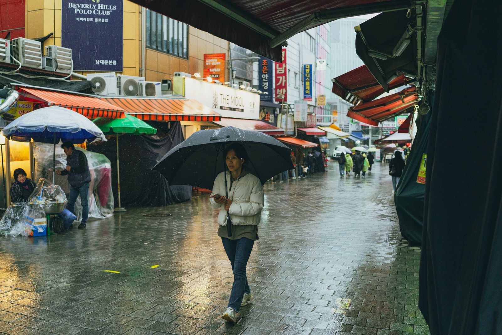 A rainy Seoul market street with umbrellas and wet pavement, representing the kind of weather that makes a long cafe reset useful.