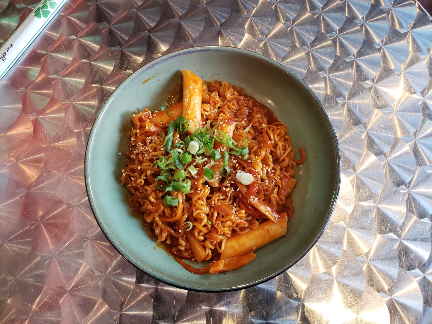 A bowl of rabokki with ramen noodles and spicy rice cakes on a metal table.