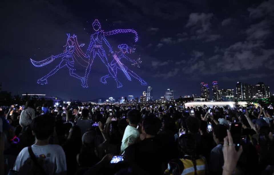 A KPop Demon Hunters drone show scene over Seoul watched by a crowd holding up phones.
