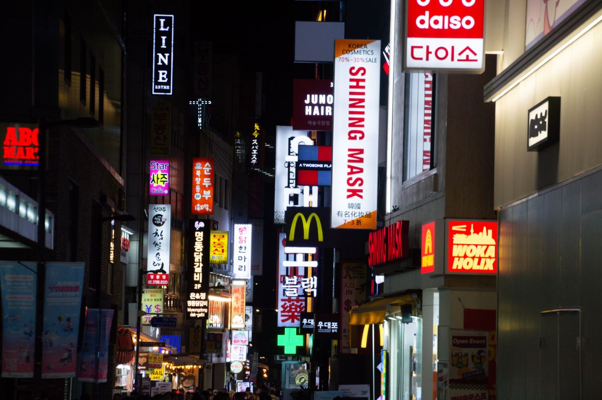 Korean cosmetics, hair, and mask shop signs glowing on a Myeongdong street at night.