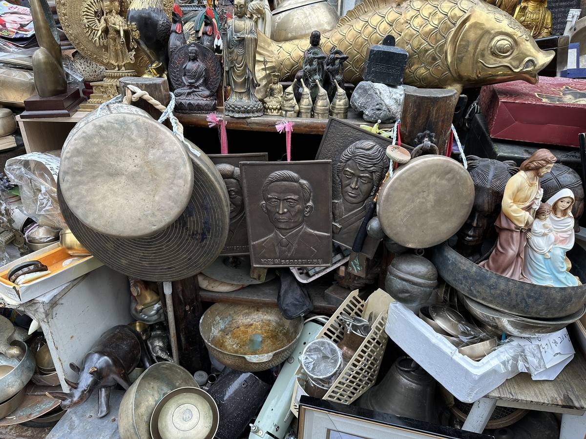 A dense unsorted pile at a Dongmyo antique stall — Buddha figures, a large brass carp, two bronze gongs, brass bowls, a Nativity figurine, and bronze presidential portrait plaques.