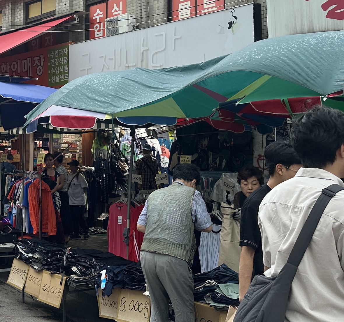 A Dongmyo stall street with handwritten price cards reading 13,000원, 10,000원, 8,000원 and 5,000원 on folded trousers, under a shop sign reading 구제나라.