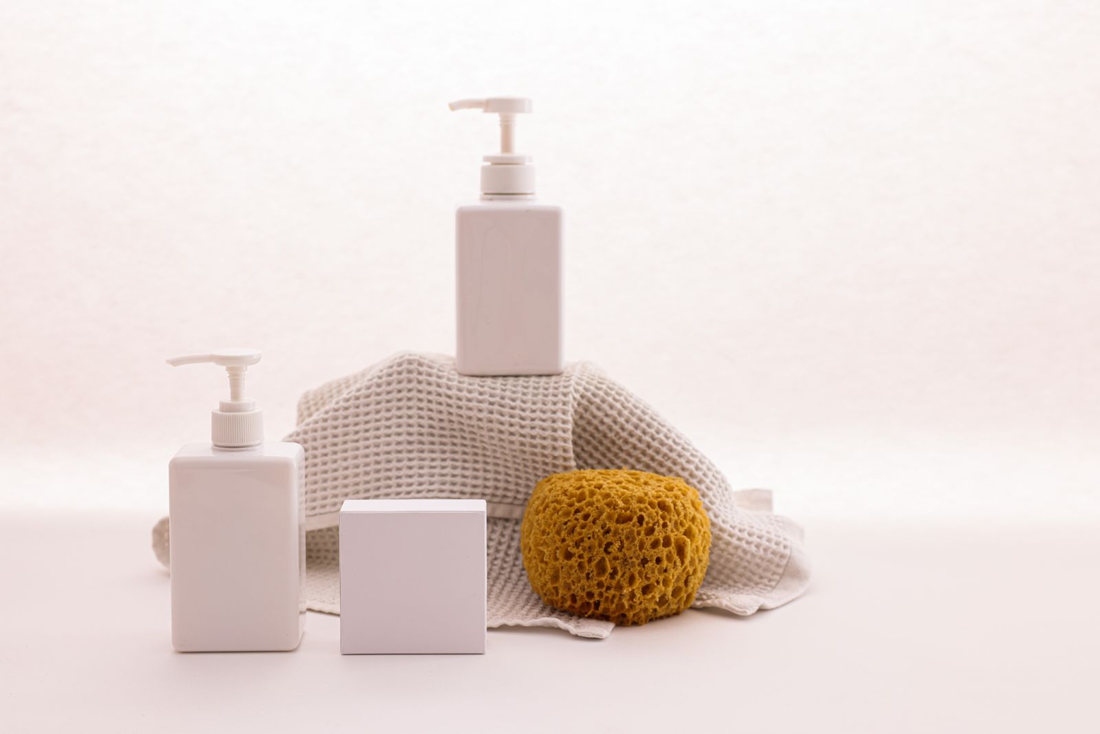 White body-care pump bottles with a sponge and towel on a soft neutral surface.