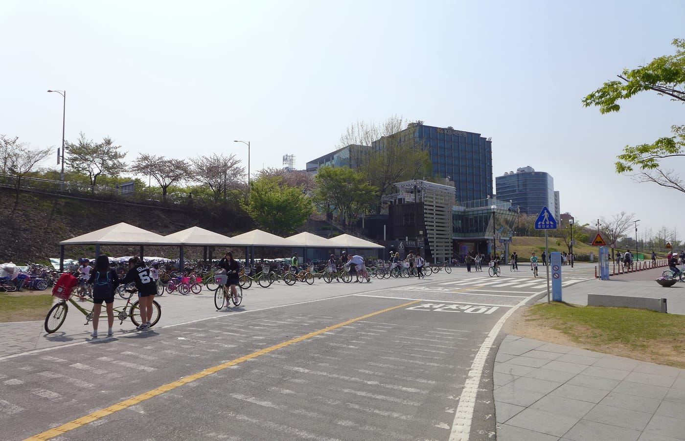 A wide paved area in a Han River park with painted lane markings and Korean text on the ground, bike-rental canopies, cyclists and walkers sharing the surface.
