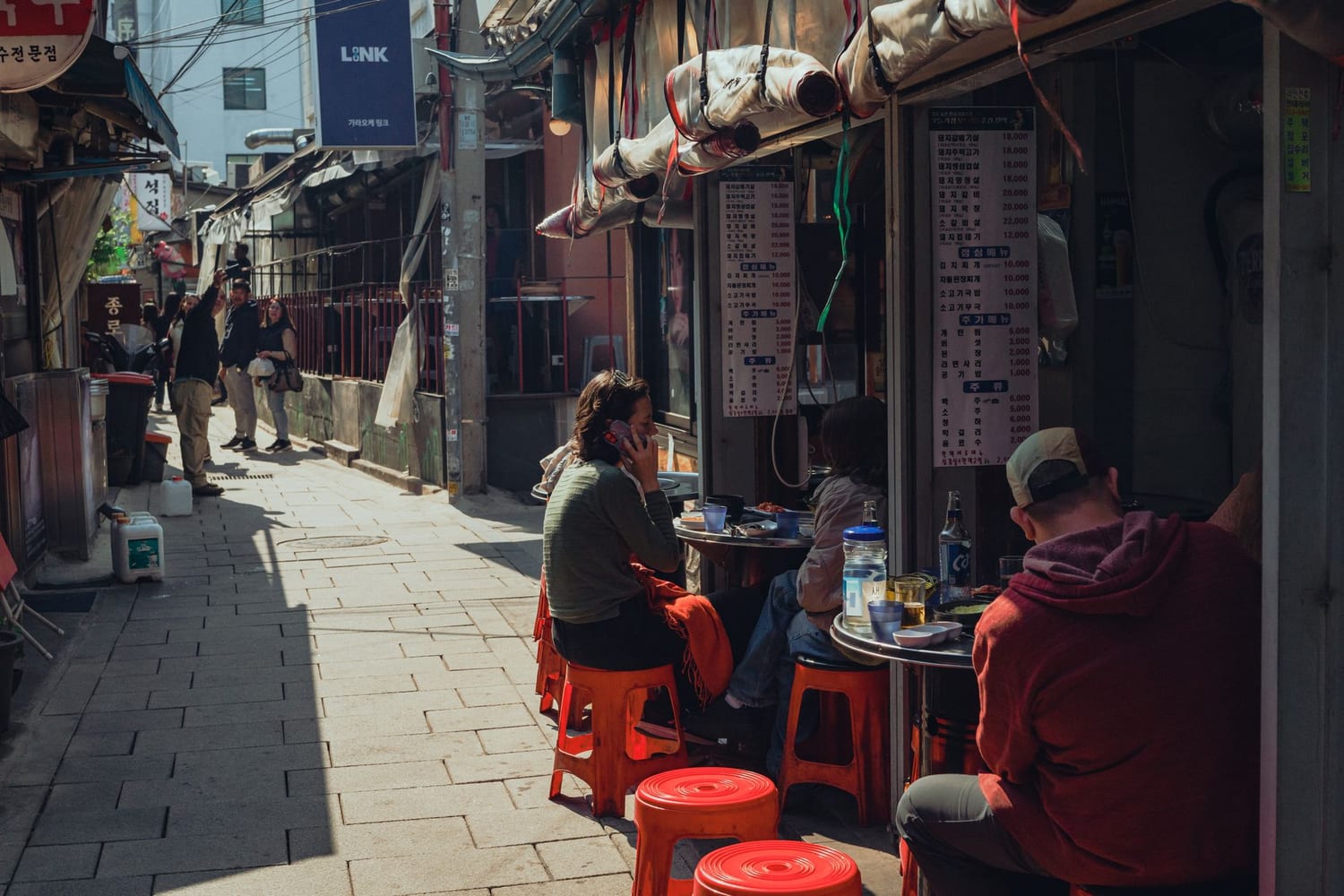 A narrow sunlit Seoul alley in Jongno with people eating at outdoor tables on red plastic stools, beside a Korean price board listing pork cuts, lunch stews and drinks.