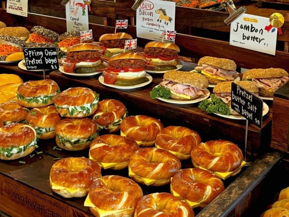 A London Bagel Museum counter at The Hyundai Seoul stacked with pretzel bagels and filled bagel sandwiches, with handwritten price cards reading 5.9 and 8.5 propped among them.