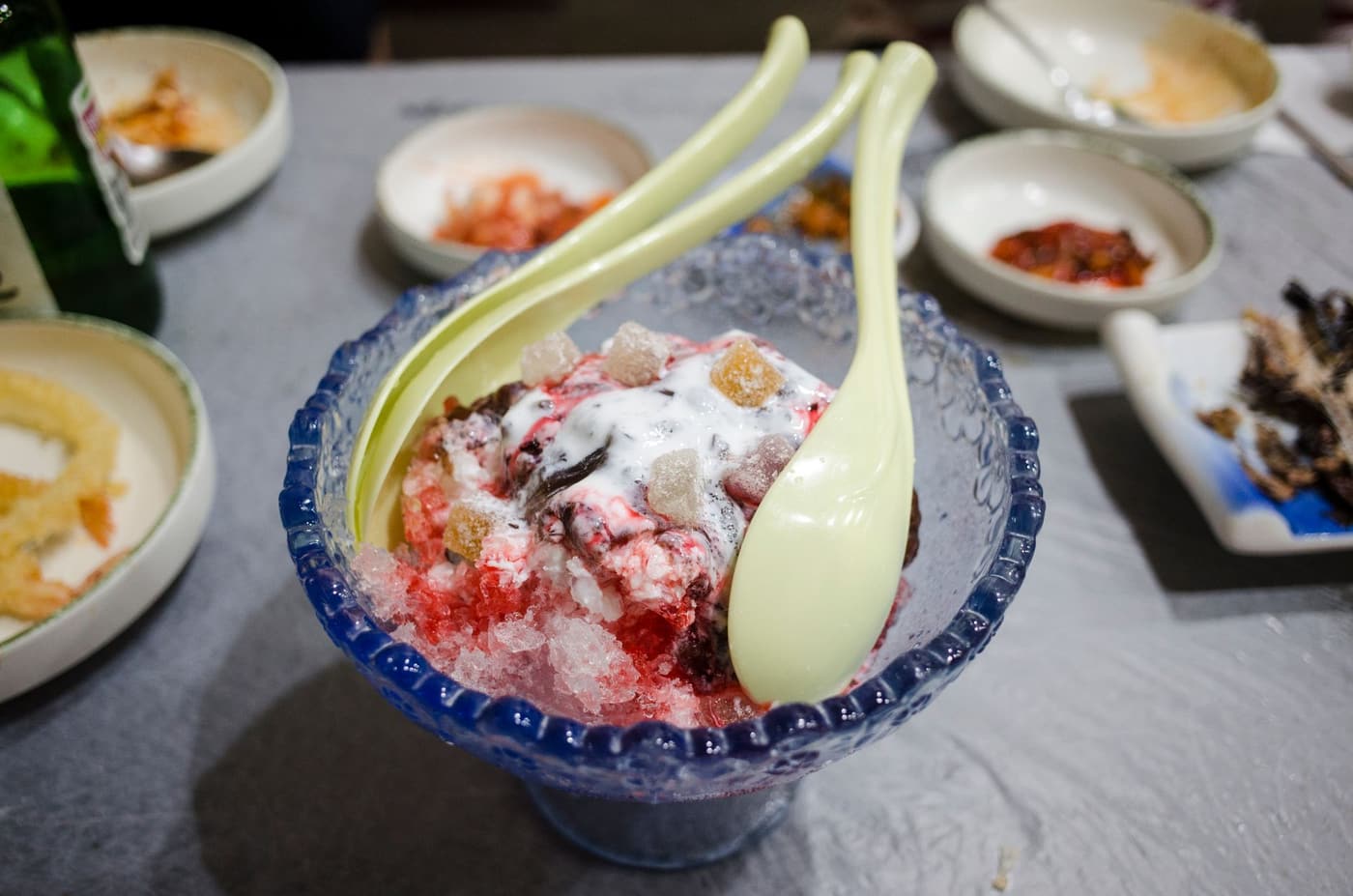 A bowl of Korean bingsu topped with fruit and syrup, two spoons resting in the shaved ice.