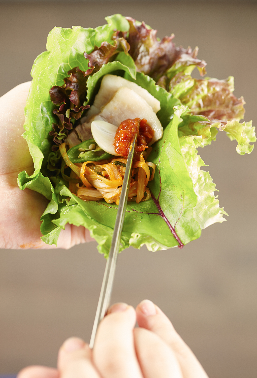 A hand holding a Korean ssam wrap with lettuce, pork, garlic, sauce, and chopsticks.