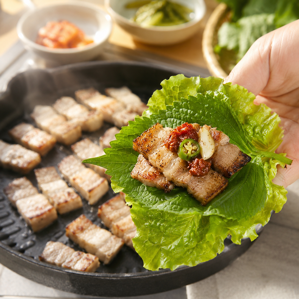 A Korean pork belly ssam wrap with lettuce, perilla, garlic, chili, and ssamjang near a grill pan.
