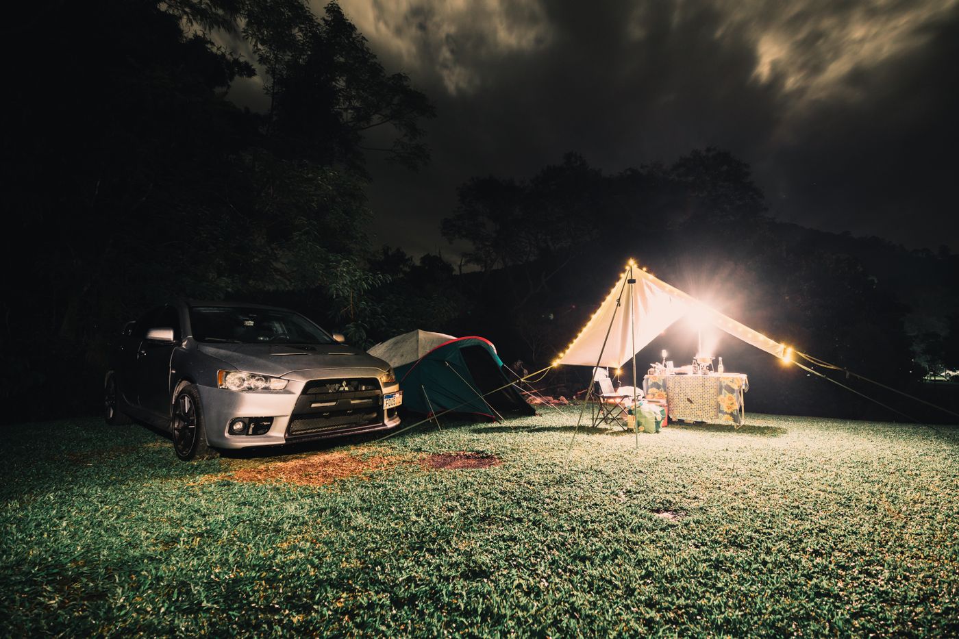 A night car-camping setup with a car, tent, lights, and table in a grassy outdoor area.