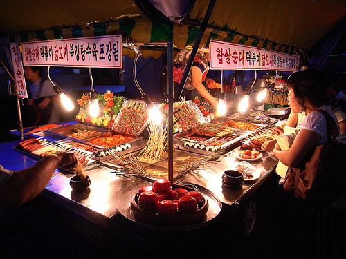 A traditional Korean pojangmacha counter with skewers, food trays, bright lamps, and the cook working within arm's reach.