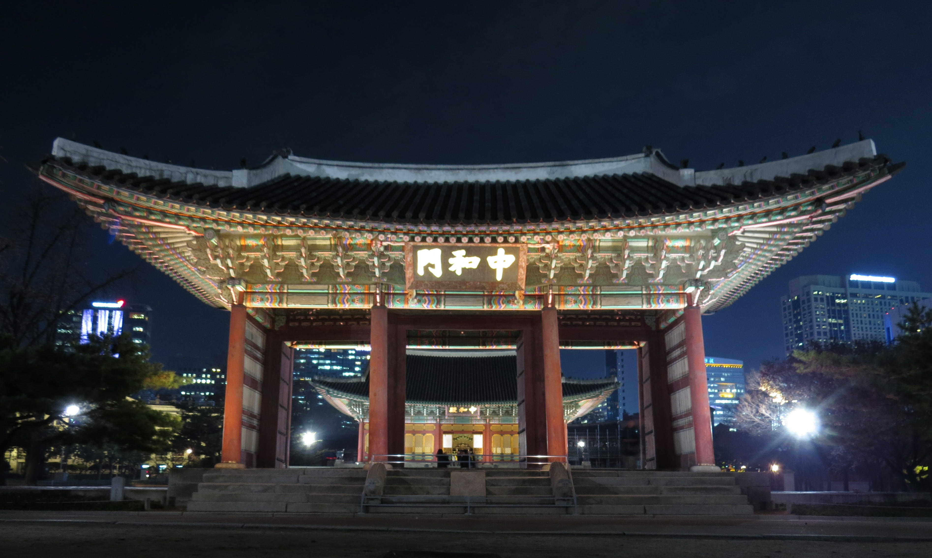 Junghwamun Gate at Deoksugung illuminated with modern Seoul buildings behind it.